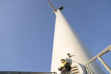 Ingenieur mit Laptop unter einer Windkraftanlage an einem sonnigen Tag - EKGF00518