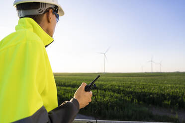 Engineer with walkie-talkie looking at wind turbines in field on sunny day - EKGF00521