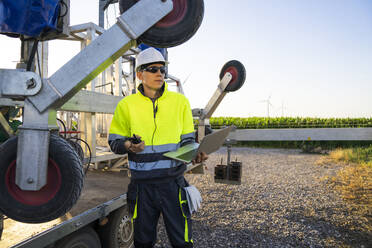 Engineer holding laptop and walkie-talkie by maintenance machine of wind turbine - EKGF00560