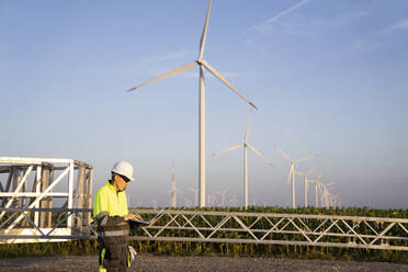 Engineer using laptop by wind turbine field in front of sky - EKGF00568