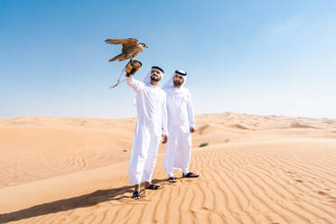 Two middle-eastern men wearing traditional emirati arab kandura bonding in the desert and holding a falcon bird - Arabian muslim friends meeting at the sand dunes in Dubai - DMDF04421