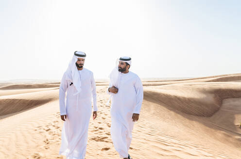 Two middle-eastern men wearing traditional emirati arab kandura bonding in the desert - Arabian muslim friends meeting at the sand dunes in Dubai - DMDF04423