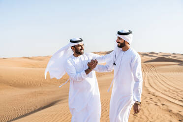 Two middle-eastern men wearing traditional emirati arab kandura bonding in the desert - Arabian muslim friends meeting at the sand dunes in Dubai - DMDF04424