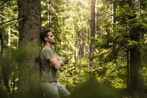 Hiker with arms crossed leaning on tree at forest - DIGF20531