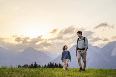 Vater und Tochter spazieren im Gras vor einer Bergkette bei Sonnenuntergang - DIGF20535