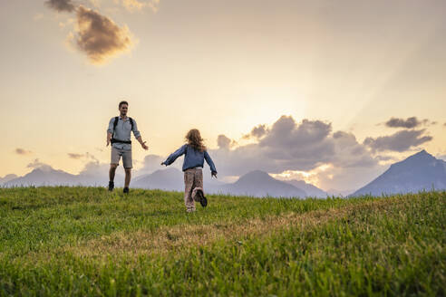 Girl running towards father standing on grass in front of mountains - DIGF20544