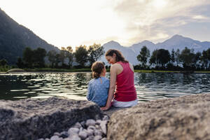 Mother and daughter sitting on rock by lake at sunset - DIGF20568