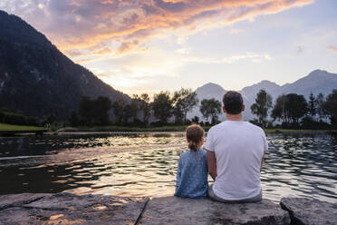 Vater und Tochter sitzen auf einem Felsen und bewundern den See bei Sonnenuntergang - DIGF20569