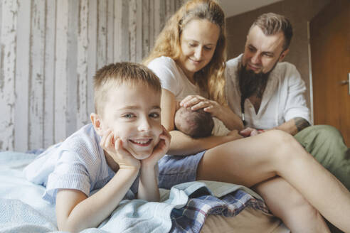 Lächelnder Junge mit Mutter, die ihre neugeborene Tochter zu Hause auf dem Bett stillt - IHF01682