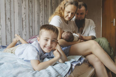 Smiling son lying next to parents with newborn daughter on bed at home - IHF01683