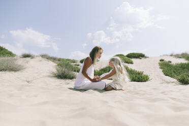 Mutter und Tochter sitzen mit gekreuzten Beinen am Strand an einem sonnigen Tag - SIF00822