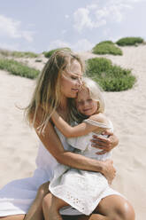 Smiling daughter sitting on mother's lap at beach on sunny day - SIF00825