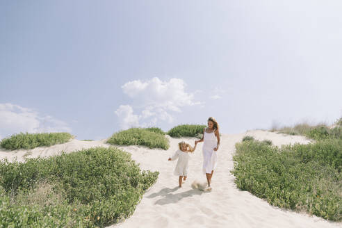 Mother and daughter holding hands and walking on sand at beach - SIF00835