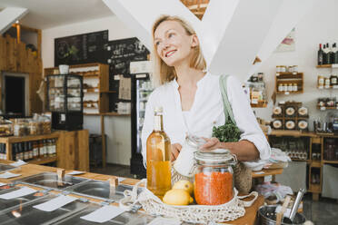 Happy woman holding jar and standing with groceries at table - NDEF01106