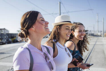 Happy friends with smart phones standing at tram tracks on sunny day - DCRF01828