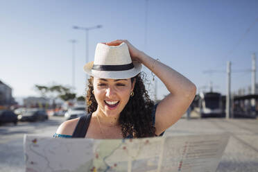 Glückliche Frau mit Hut, die an einem sonnigen Tag eine Karte betrachtet - DCRF01847