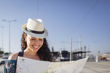 Lächelnde Frau mit Hut, die an einem sonnigen Tag eine Karte betrachtet - DCRF01848