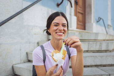 Glückliche Frau sitzt mit Eiswaffel auf einer Treppe - DCRF01855