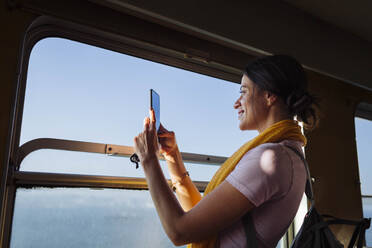 Smiling young woman photographing through smart phone in train - DCRF01875