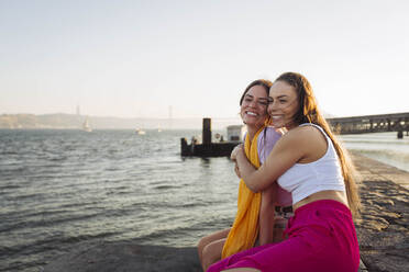 Happy woman hugging friend sitting on promenade - DCRF01901