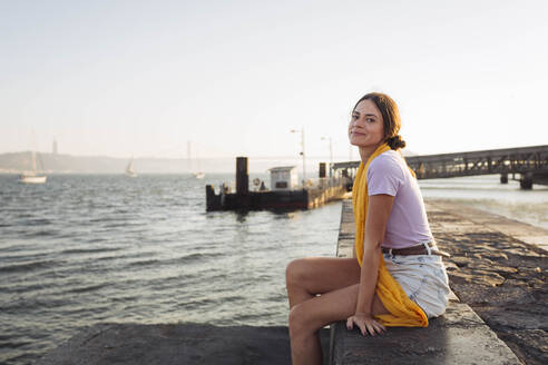 Smiling young woman sitting on promenade - DCRF01902