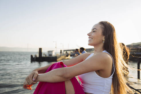 Happy woman with long hair sitting at promenade on sunny day - DCRF01908