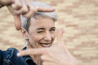 Lächelnde Frau zwinkert und zeigt Fingerrahmen-Zeichen vor einer Wand - JCZF01266