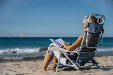Ältere Frau sitzt auf einem Liegestuhl und liest ein Buch am Strand - GDBF00084