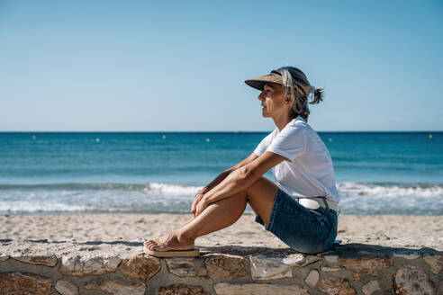 Contemplative woman sitting on promenade at beach - GDBF00086