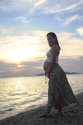 Schwangere Frau lächelnd und stehend am Strand unter bewölktem Himmel - DMHF00153