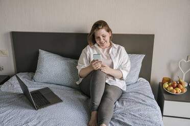 Happy freelancer using smart phone near laptop on bed at home - YBF00209