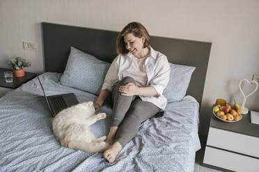 Lächelnde Frau spielt mit Katze in der Nähe von Laptop auf Bett zu Hause - YBF00210