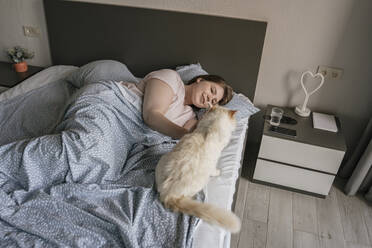 Glückliche Frau entspannt sich mit Katze auf dem Bett zu Hause - YBF00213