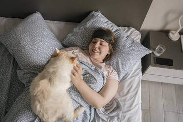 Lächelnde Frau mit Augenmaske streichelt Katze auf dem Bett - YBF00220