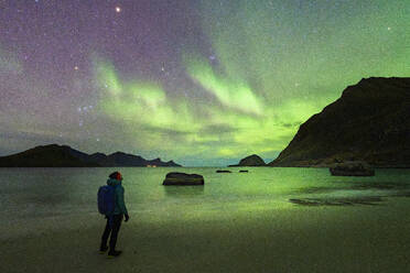 Man with backpack admiring the bright green lights of Aurora Borealis (Northern Lights) from Haukland beach, Lofoten Islands, Nordland, Norway, Scandinavia, Europe - RHPLF27357