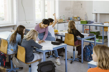 Female teacher assisting schoolgirls while sitting on desk in classroom - MASF38296