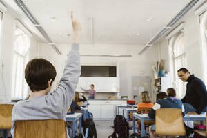 Rear view of schoolboy raising hand while attending lecture in classroom - MASF38297
