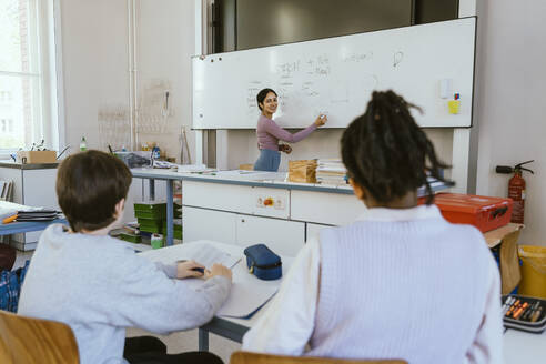 Smiling female teacher writing on whiteboard while teaching male students sitting in classroom - MASF38300