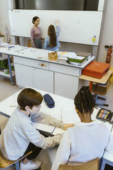 High angle view of boy helping male friend while sitting on desk in classroom - MASF38302