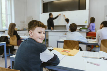 Porträt eines lächelnden Jungen, der über die Schulter schaut, während er auf dem Schreibtisch im Klassenzimmer sitzt - MASF38303