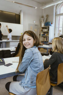 Seitenansicht Porträt eines lächelnden Mädchens, das eine Jeansjacke trägt und auf einem Stuhl im Klassenzimmer sitzt - MASF38304