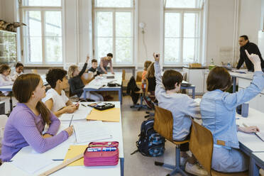 Male and female students raising hands while sitting on chairs in classroom - MASF38310
