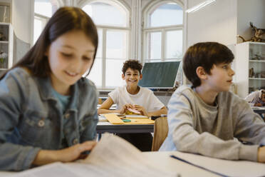 Portrait of happy schoolboy sitting at desk with friends in classroom - MASF38313