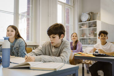 Schoolboy writing in book while sitting at desk with friends in classroom - MASF38314