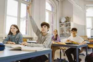 Junge hebt die Hand, während er mit seiner Freundin am Schreibtisch im Klassenzimmer sitzt - MASF38315