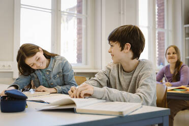 Happy boy laughing while female friend sitting at desk in classroom - MASF38316