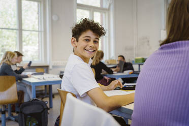 Seitenansicht Porträt eines lächelnden Schuljungen sitzt am Schreibtisch im Klassenzimmer - MASF38320