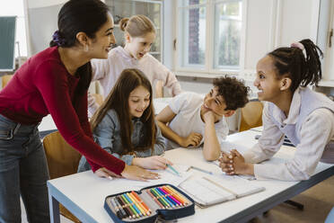 Happy female teacher leaning at desk with students in classroom - MASF38326