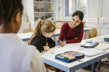 Smiling female teacher explaining schoolgirl sitting at desk in classroom - MASF38330