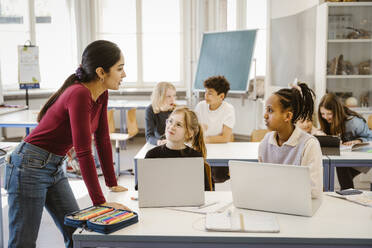 Female teacher talking to schoolgirls sitting with laptops on desk in classroom - MASF38333